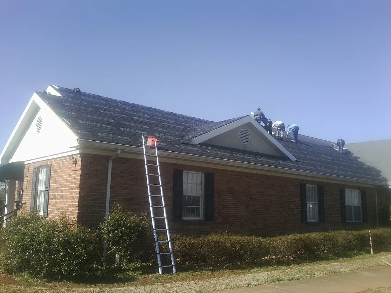 Crew tearing shingles off a brick ranch roof with a ladder set — Emergency Roof Repair work by our crew in Oelwein