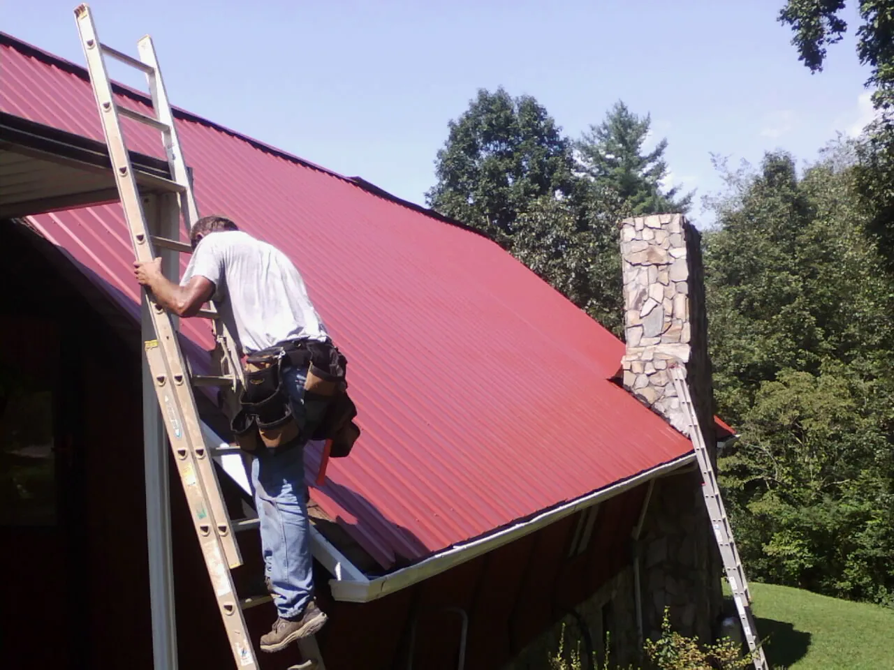 Roofer on a ladder beside a red metal roof and stone chimney — Soffit & Fascia Repair by Roofing Brothers in Oelwein